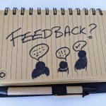 On a recycled, brown notebook is scribbled the word "Feedback?", floating over three stick people each with speech bubbles