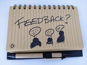 On a recycled, brown notebook is scribbled the word "Feedback?", floating over three stick people each with speech bubbles